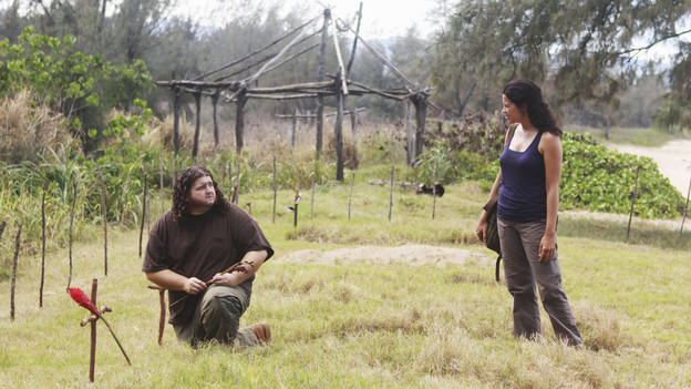 Screencap from LOST: Hurley is crouched on the ground next to a grave on the island, looking up and talking to Ilana.